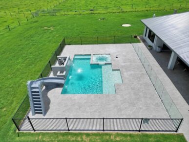Wide view of a fenced Saint Remy pool installation with spa, slide, and surrounding green rural landscape